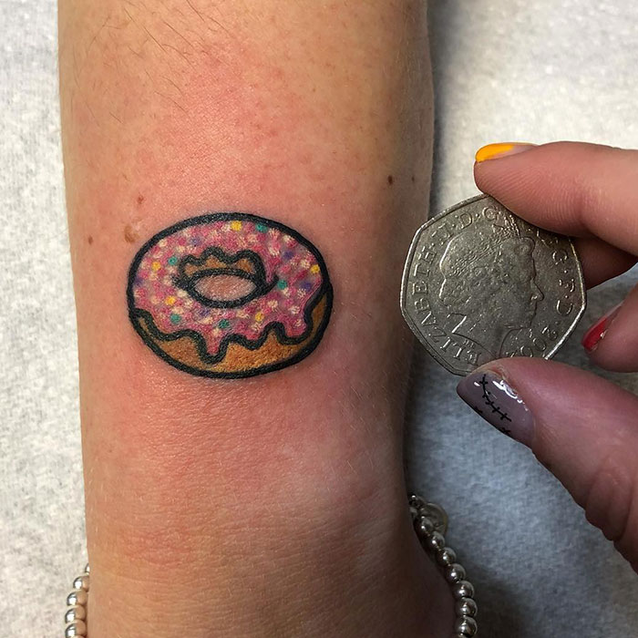 A small tattoo of a pink frosted donut with sprinkles, playfully detailed and shown next to a coin for size comparison.