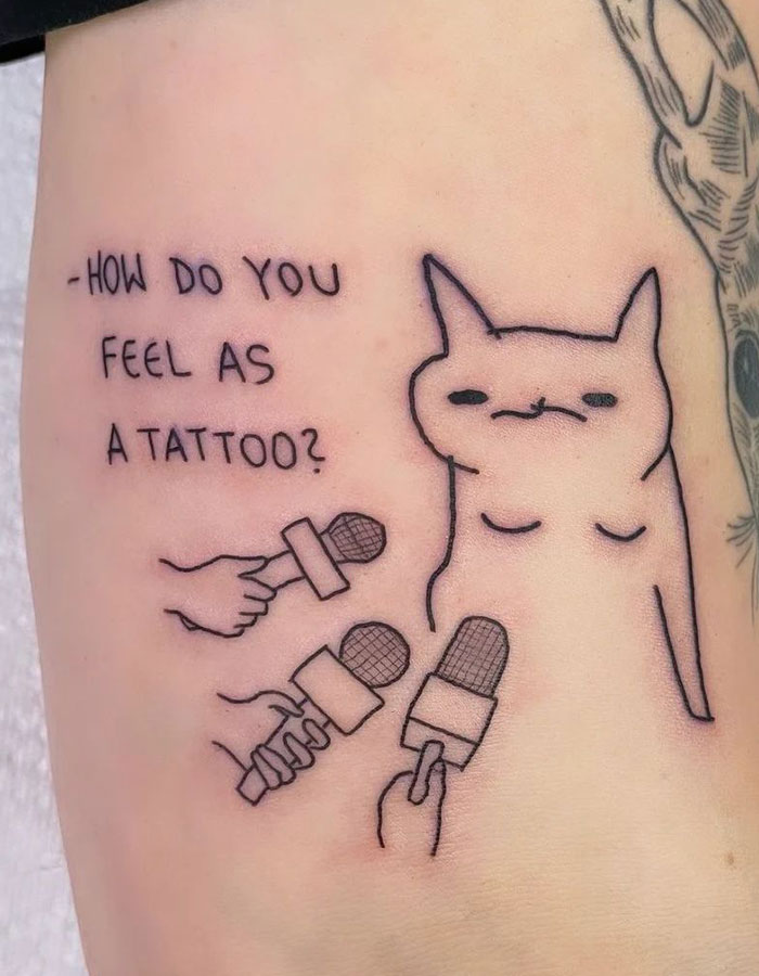 A small tattoo featuring a humorous drawing of a cat being interviewed by multiple microphones, with the text, "How do you feel as a tattoo?"