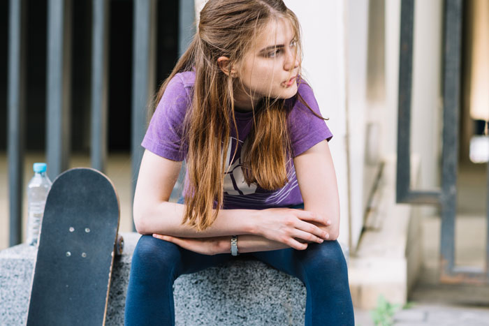 Teen girl sitting on a ledge, wearing a purple shirt, near a skateboard, looking contemplative.