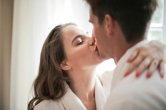 A woman kissing a man in a close embrace, both in a soft-lit room, symbolizing a complex affair.