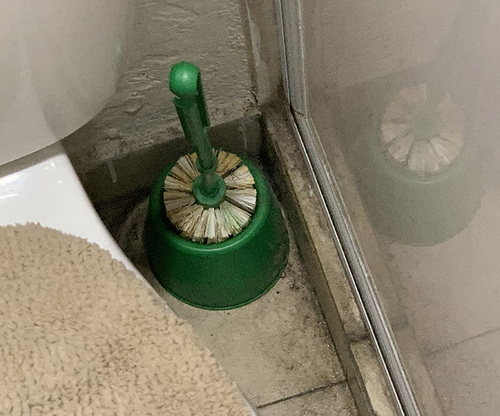 Dirty bathroom corner with a used toilet brush and holder. Girlfriend lives filthy, boyfriend unaware how to tell her.