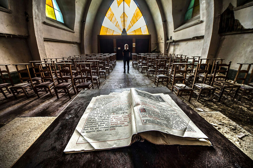 Abandoned sacred place in Europe with open book on decaying pew, stained glass windows, and lone figure in the distance.