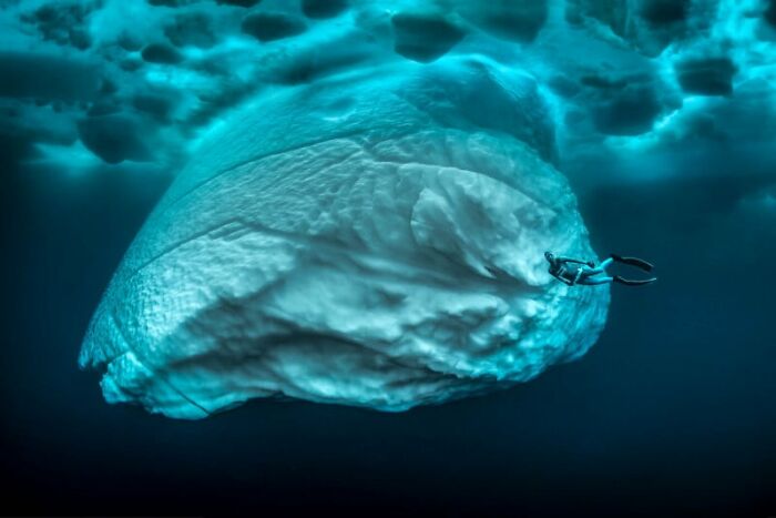 Diver swimming under a massive iceberg, showcasing breathtaking photography focused on sustainability.