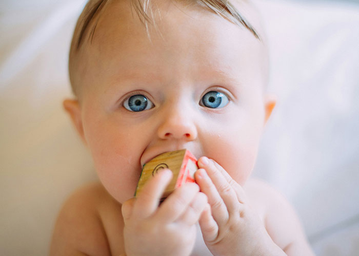 Baby with blue eyes chewing on a wooden block, representing childhood innocence amidst social challenges.