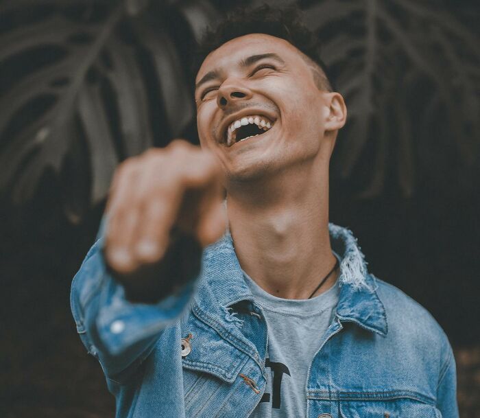 A man in a denim jacket laughing and pointing, reminded of the funniest joke he's been told.