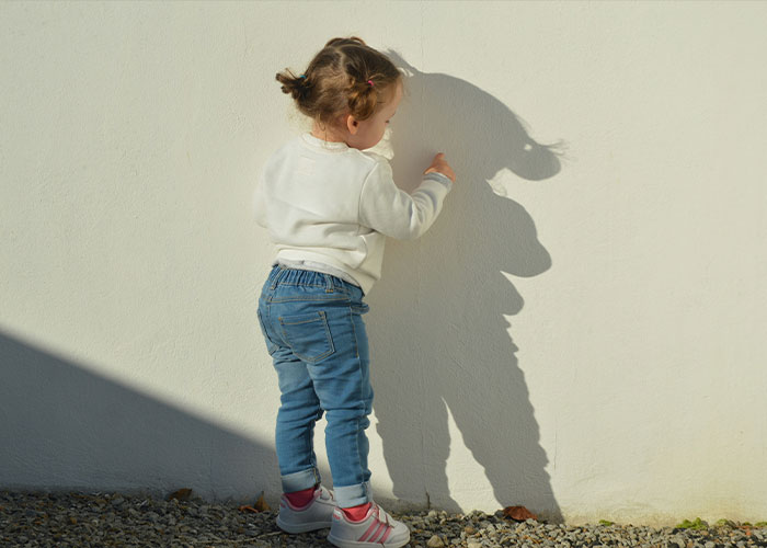 Toddler girl playing with her shadow on a sunny day, dressed in jeans and a white sweater.