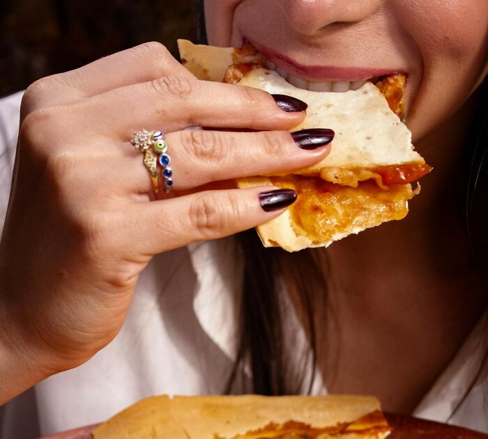 Person eating pizza, wearing a ring with navy nails, pondering a funny joke they still think about to this day.