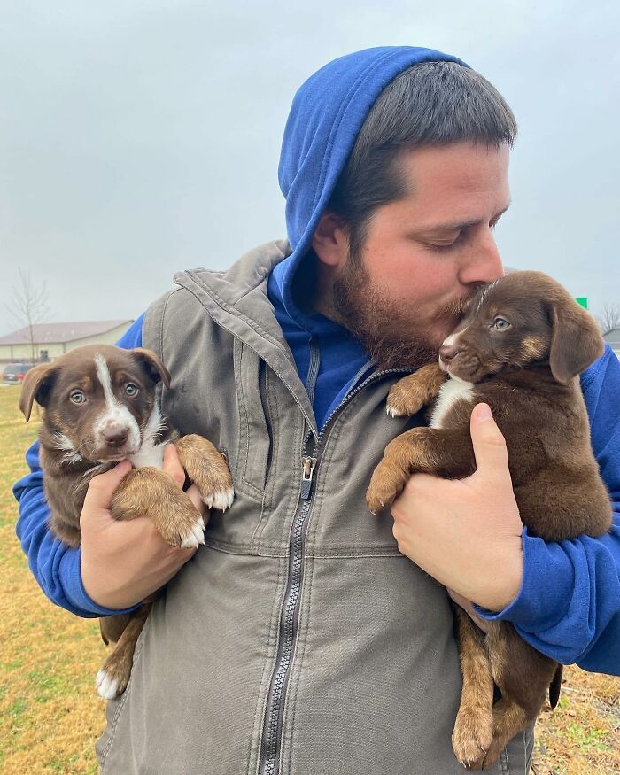 Man holding and kissing two rescued puppies, dressed in a blue hoodie, outdoors.