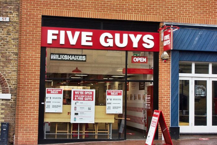 Front view of Five Guys restaurant, known for its milkshakes, with red signage and brick facade, open for customers.