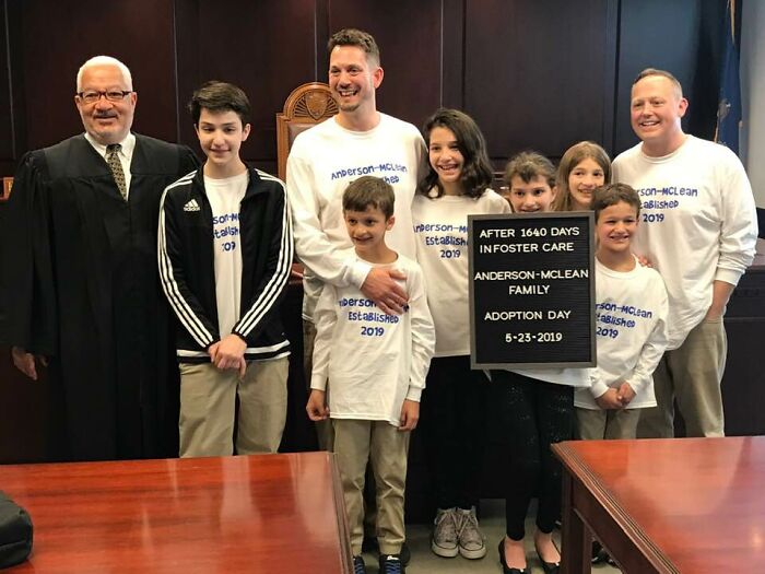 Family celebrates adoption day with judge, holding sign after foster care, embodying wholesome adoption stories.