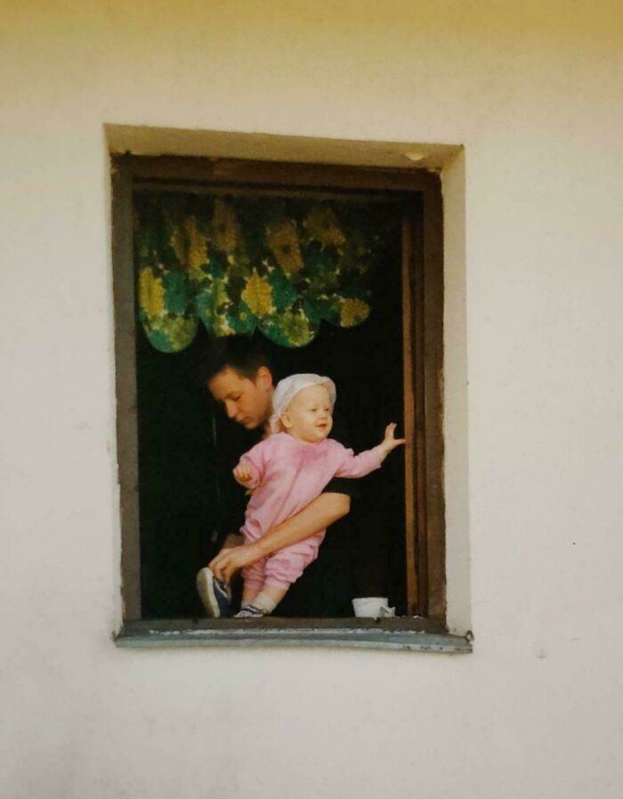 Child in pink outfit held by adult through window, resembling a Renaissance painting.