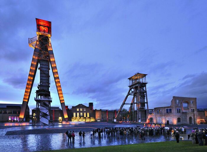 People gather at dusk near a converted industrial building with illuminated towers, showcasing one of the best building conversions.