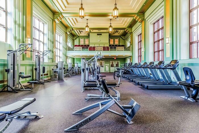 Spacious gym with treadmills and fitness equipment in a converted building featuring vintage architecture and large windows.