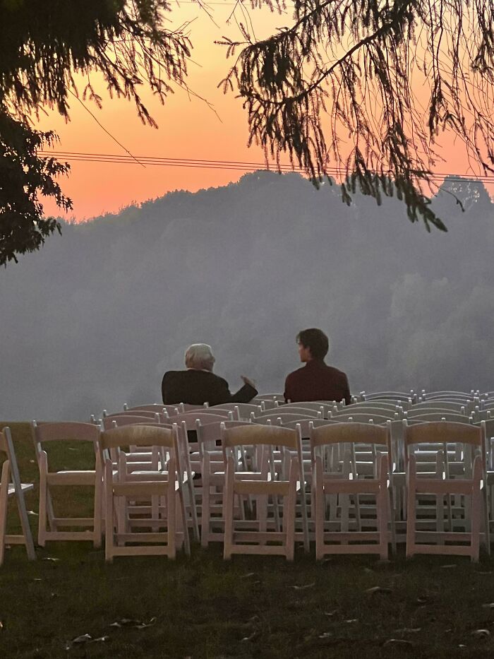 Two people seated among empty chairs at sunset, evoking the style of Renaissance paintings.