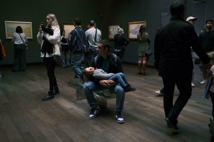 Man sitting with child in a museum, scene resembling Renaissance painting, surrounded by other visitors and framed artwork.