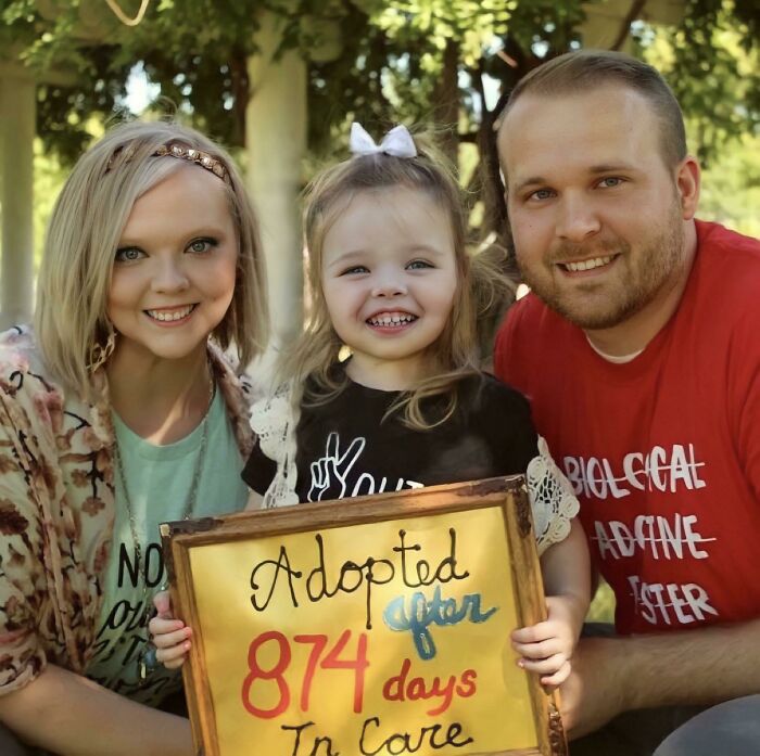 Smiling family with a sign reading "Adopted after 874 days in care," showcasing a joyful adoption story.