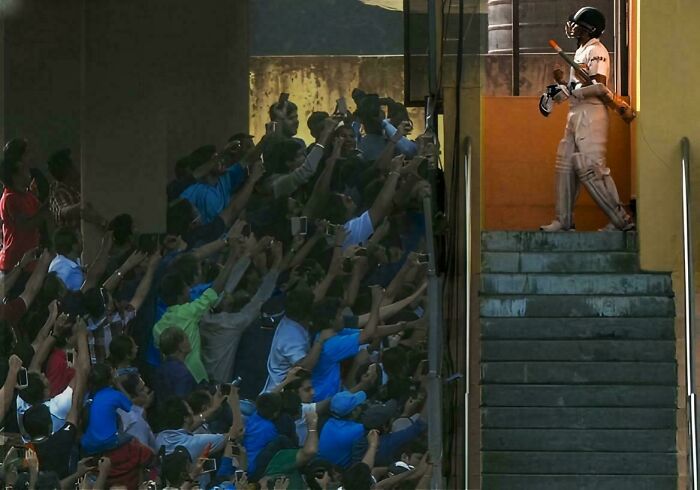 Cricketer emerging from stairs, with crowd resembling a Renaissance painting, capturing the moment with phones.