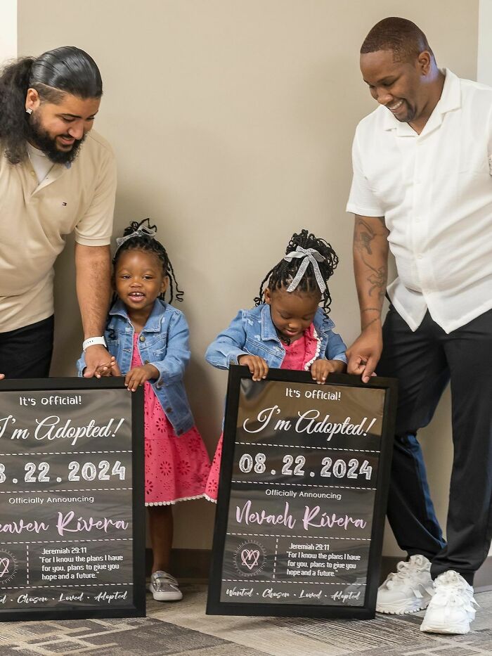 Two smiling parents with adopted daughters holding announcement signs, celebrating their wholesome adoption story.