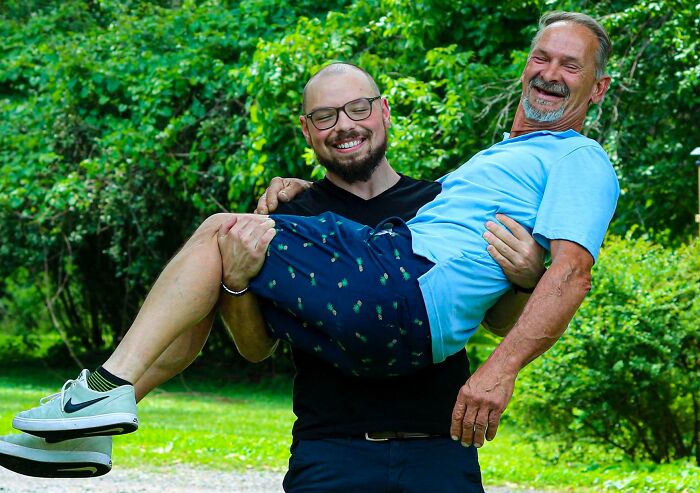 Two men smiling, one being playfully carried, showcasing a wholesome adoption story in a lush green park setting.