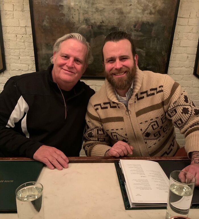 Two men in a cozy restaurant setting, smiling at the camera, illustrating a heartfelt moment related to adoption stories.