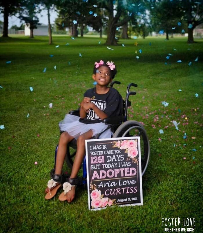 A young girl in a wheelchair celebrates adoption with a decorated sign in a park setting.