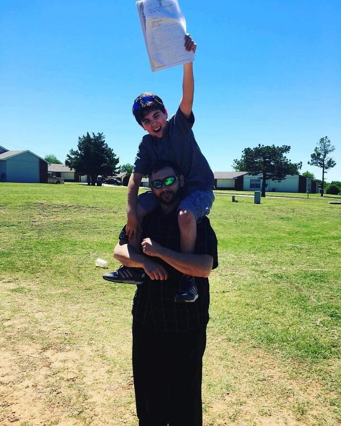 Child sitting on a man's shoulders holding up adoption papers, celebrating a wholesome moment outdoors.