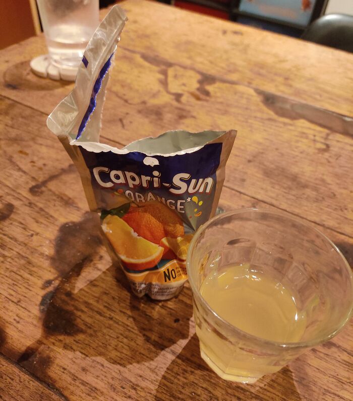 Capri-Sun pouch opened incorrectly by boyfriend or husband, next to a glass of juice on a wooden table.