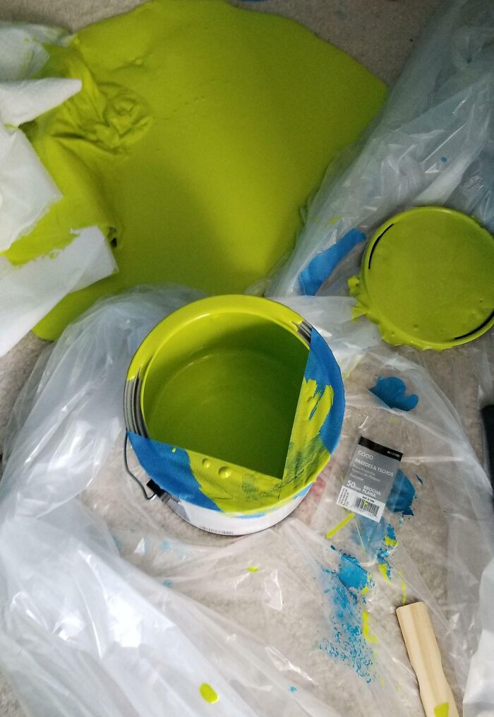 Paint spill on the floor with open lime green paint can, plastic sheets, and scattered blue paint, showcasing a messy accident.