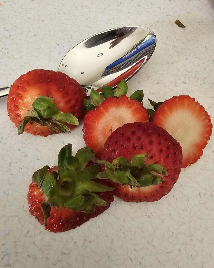 Strawberries with tops left on, sliced in a frustrating manner, beside a reflective spoon.