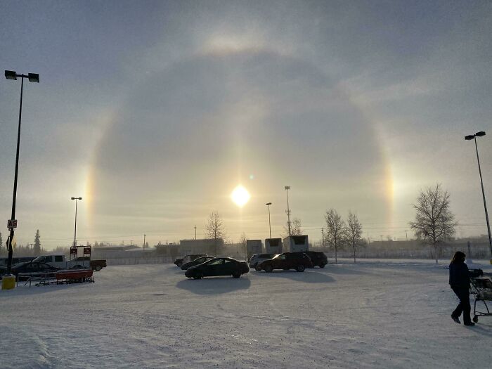 Ice-cold winter scene with a halo around the sun, snow-covered parking lot, and a person pushing a cart.