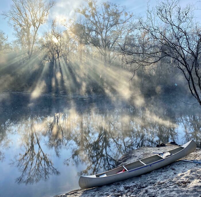 Canoe by a misty river in winter, surrounded by bare trees and soft sunlight.