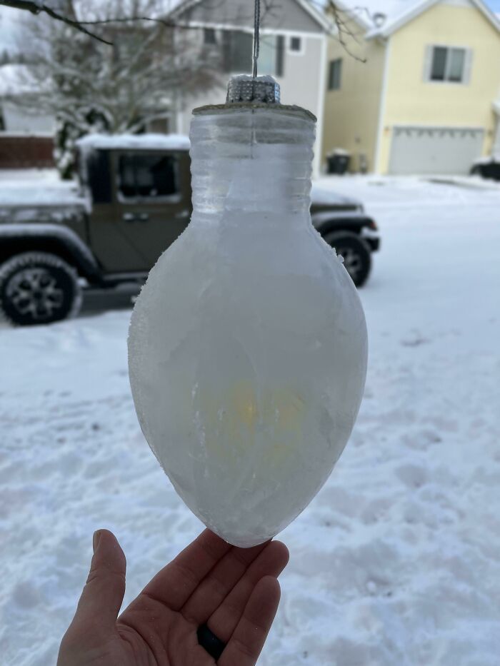 Frozen light bulb hanging outside, surrounded by snow-covered houses and cars, capturing ice-cold winter scenery.