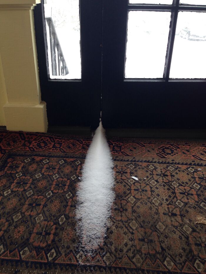 Snow entering through a gap under a dark door onto a patterned rug, highlighting ice-cold winter conditions.