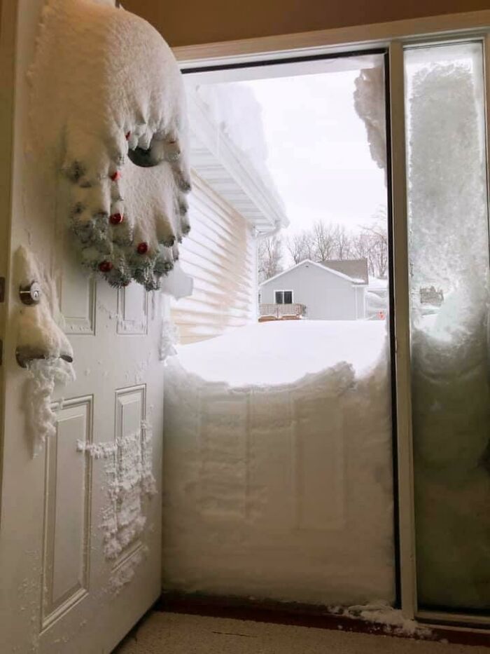 Snow piled against an open door, revealing a wreath covered in ice, capturing a winter scene.