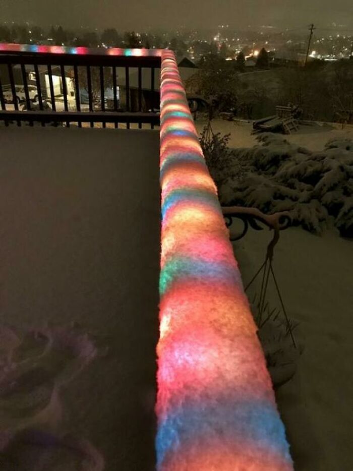 Colorful lights on snow-covered railing during ice-cold winter evening, overlooking a snowy town.