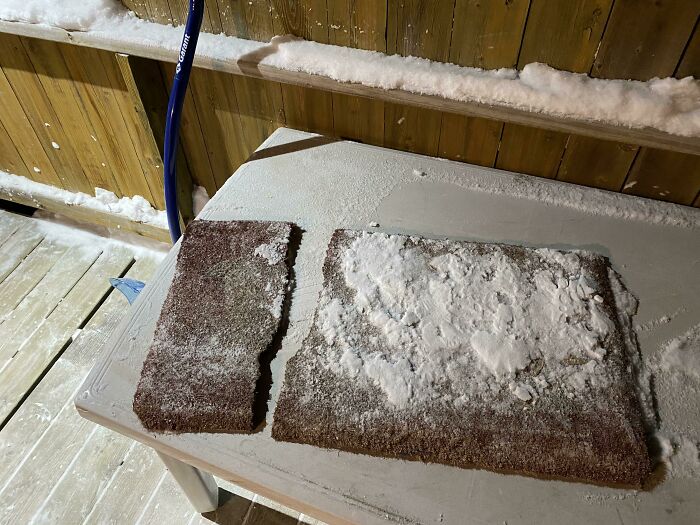 Snow-covered doormats on a wintery outdoor table with a snow shovel leaning against a wooden railing.