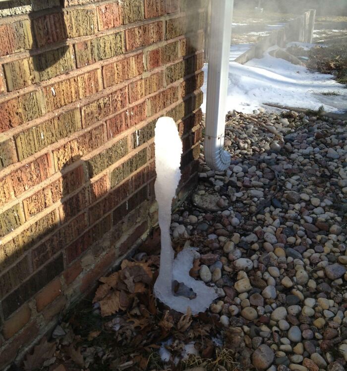 Icicle formed near a brick wall and gutter in a winter scene, showcasing ice-cold weather conditions.