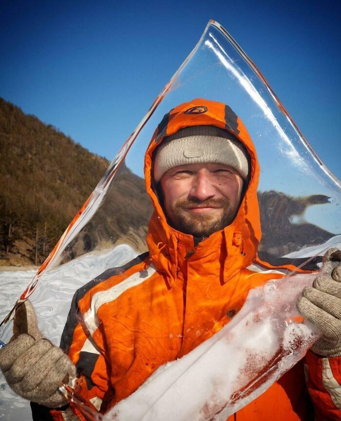 Man in orange jacket holds a triangular ice chunk against a clear blue winter sky. Ice-Cold-Winter-Pictures scene.