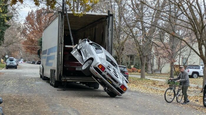 A silver car hangs off a truck, appearing to be an expensive unfortunate incident on a residential street.