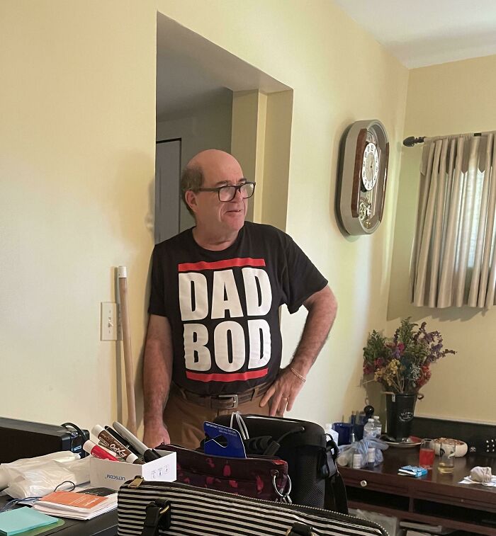 Man wearing a "Dad Bod" t-shirt, standing in a living room, humorously embodying hilarious in-laws theme.