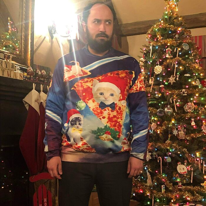 Man in a colorful cat and pizza sweater stands by a decorated Christmas tree, showcasing hilarious in-laws humor.