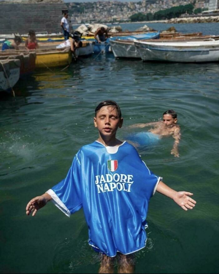 Boy in a blue "J'adore Napoli" shirt standing in the water, resembling a scene from a Renaissance painting.