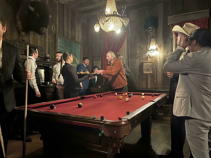 People around a pool table in dimly lit room, resembling a renaissance painting scene with rich detail and dramatic lighting.