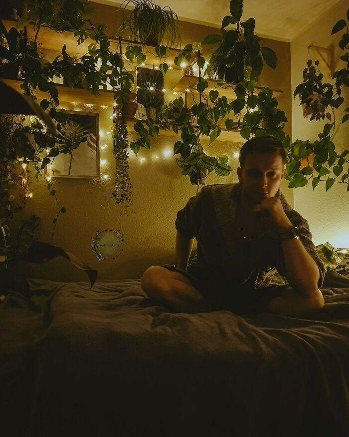 Cozy room with lush indoor garden plants and ambient string lights, person sitting thoughtfully on the bed.