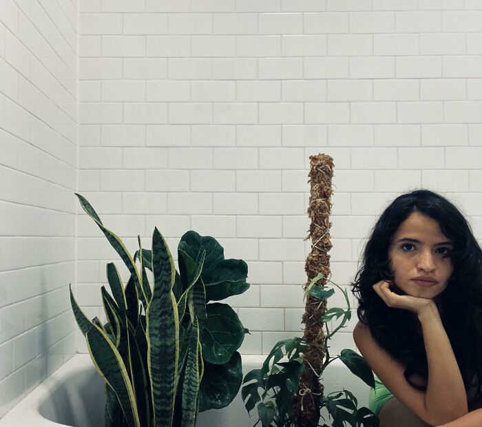 Person sitting with indoor garden plants in a white-tiled room.