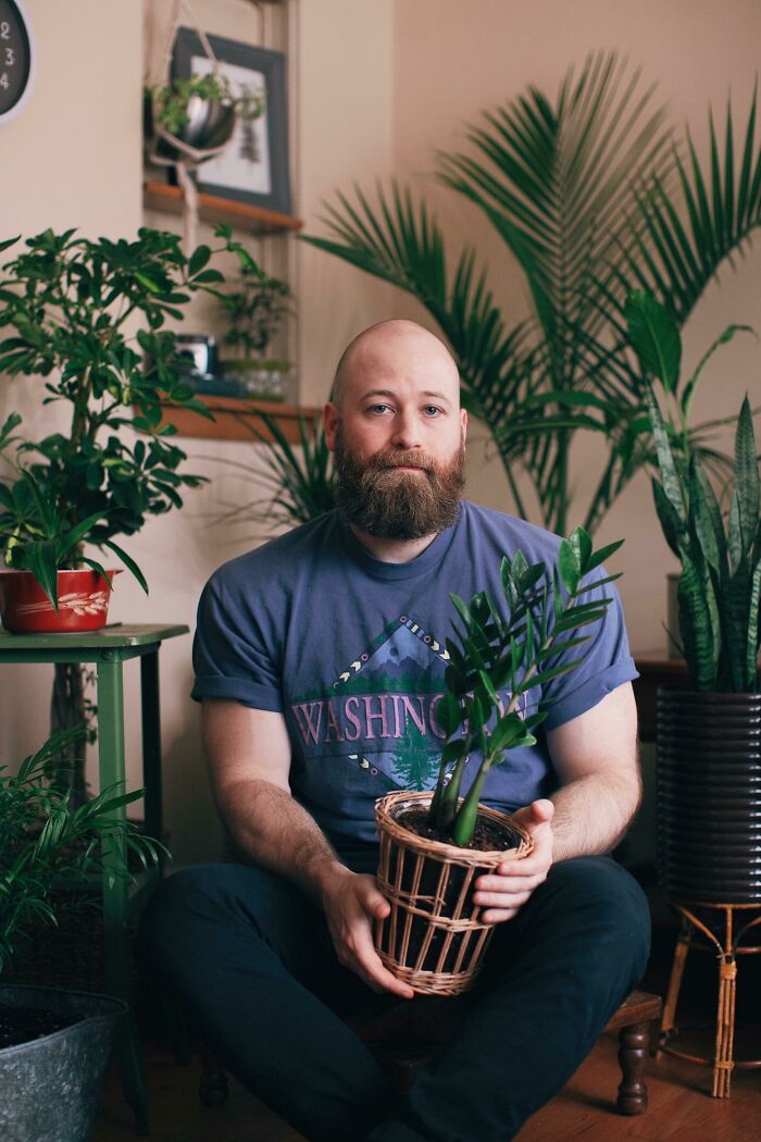 Man sitting indoors with various garden plants, holding a potted plant.