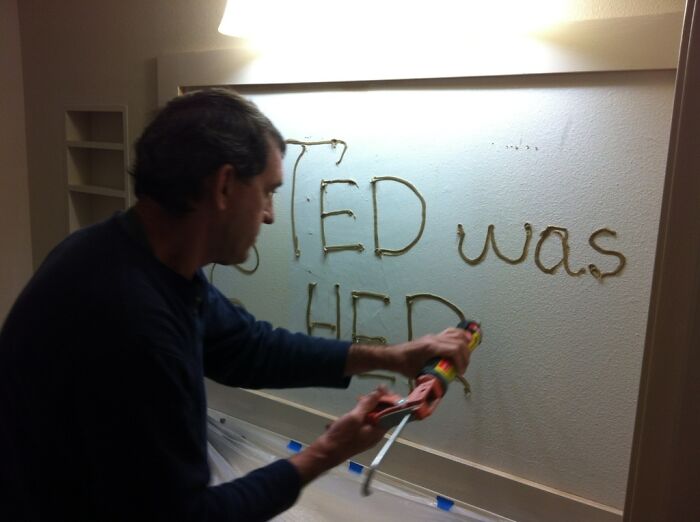 Man using caulking gun to humorously write on wall, showcasing hilarious in-laws moment.