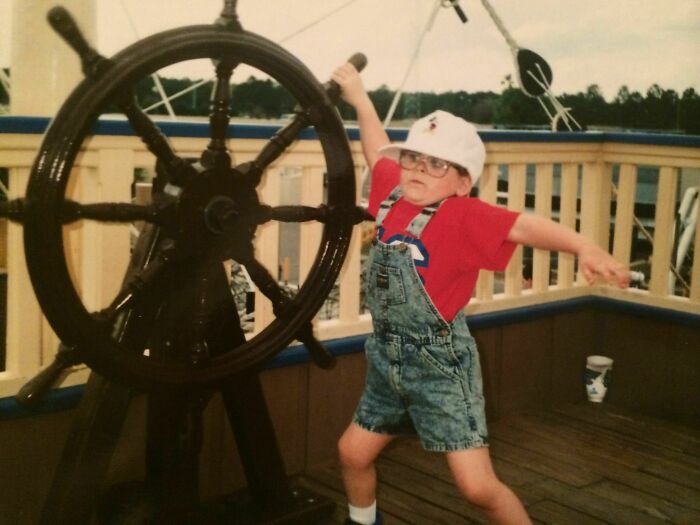 Child in overalls and a cap humorously steering a ship's wheel, embracing comedic adventure.