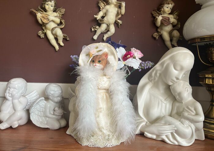 Cat figurine in a vintage outfit with fluffy boa, surrounded by angel sculptures, showcases a humorous in-laws gift idea.