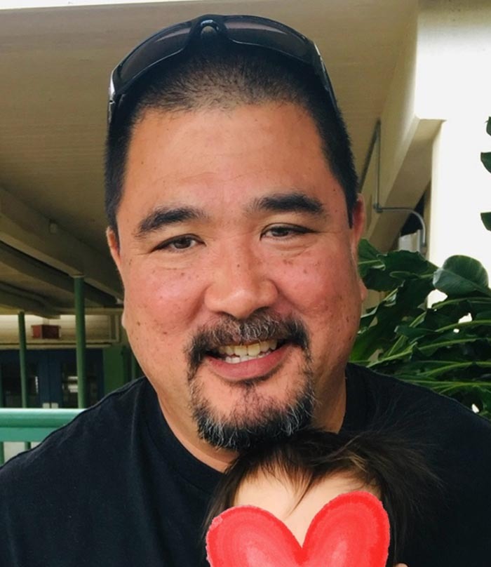 Hannah Kobayashi's recently deceased father Ryan, pictured with sunglasses on head, standing indoors near green plants.